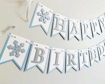 May include: A blue and white banner with silver glitter letters that reads "Happy Birthday". The banner has snowflake cutouts on each end.