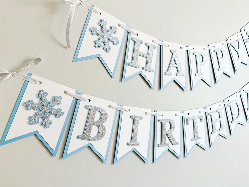 May include: A blue and white banner with silver glitter letters that reads "Happy Birthday". The banner has snowflake cutouts on each end.