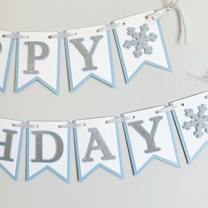 May include: A blue and white banner with silver glitter letters that reads "Happy Birthday" with a snowflake on each end.