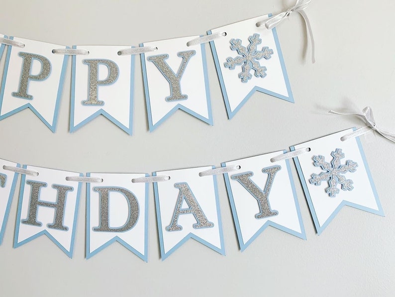 May include: A blue and white banner with silver glitter letters that reads "Happy Birthday" with a snowflake on each end.