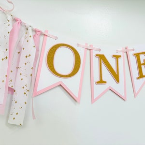 May include: Pink and gold "ONE" banner with white flags and gold glitter letters. The banner has pink and white fabric streamers attached.