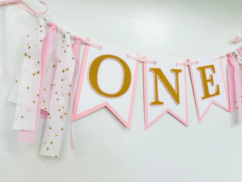 May include: Pink and gold "ONE" banner with white flags and gold glitter letters. The banner has pink and white fabric streamers attached.