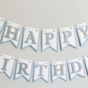 May include: A white and blue banner with silver glitter letters that reads "Happy Birthday". The banner has snowflakes on each end.