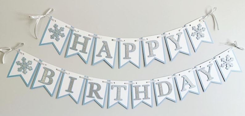 May include: A white and blue banner with silver glitter letters that reads "Happy Birthday". The banner has snowflakes on each end.