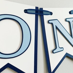 Light blue tractor first birthday ONE banner. The base pennant flags are navy blue. The letters are light blue and outlined in navy blue. The tractors have closed cabs and are light blue with navy blue tires, white wheels, white window and light orange exhaust. The tractor banner is strung on navy satin ribbon.
