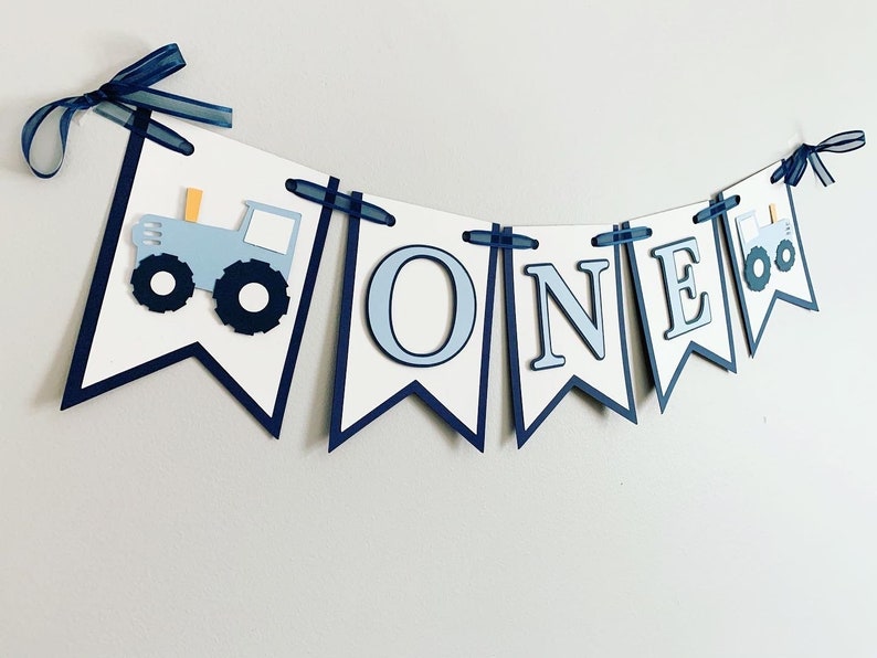 Light blue tractor first birthday ONE banner. The base pennant flags are navy blue. The letters are light blue and outlined in navy blue. The tractors have closed cabs and are light blue with navy blue tires, white wheels, white window and light orange exhaust. The tractor banner is strung on navy satin ribbon.