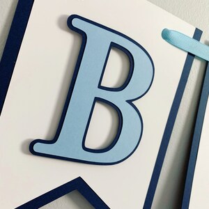 May include: A blue and white banner with the letter "B" in light blue with a dark blue outline. The banner is made of cardstock and has a navy blue border.