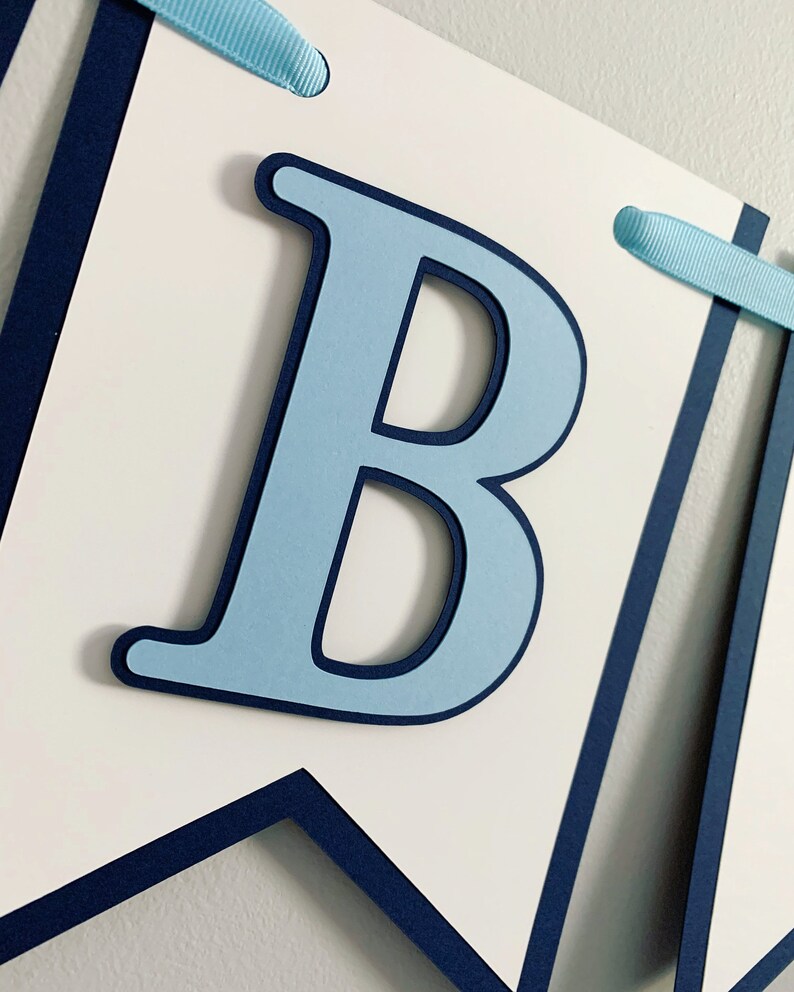 May include: A blue and white banner with the letter "B" in light blue with a dark blue outline. The banner is made of cardstock and has a navy blue border.