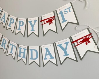 May include: A blue and gray banner with the words "Happy 1st Birthday" in blue letters. The banner has red airplane cutouts.