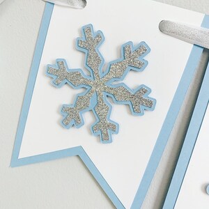 May include: A white banner with a light blue border and a silver glitter snowflake in the center. The banner has a hole punched in the top corner for hanging.