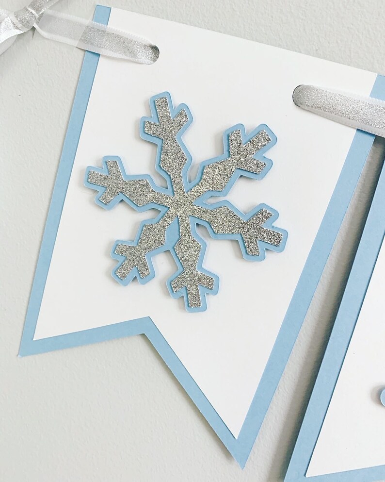 May include: A white banner with a light blue border and a silver glitter snowflake in the center. The banner has a hole punched in the top corner for hanging.