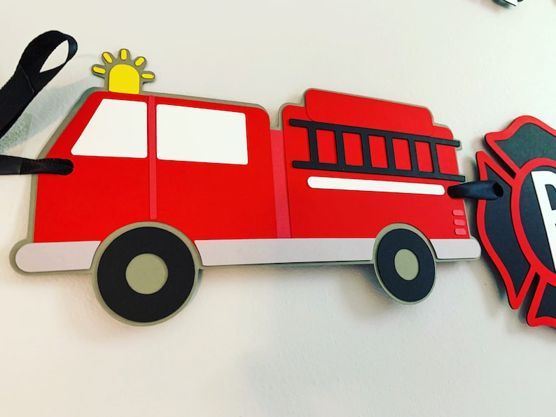 May include: Red fire engine paper cutout with a black ladder, white windows, and black wheels. The engine has a yellow star on top. The cutout is part of a fire engine themed banner.