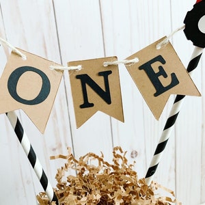 May include: A brown paper banner with the word "ONE" in black letters, attached to black and white striped straws with red and black tractor wheels on top.
