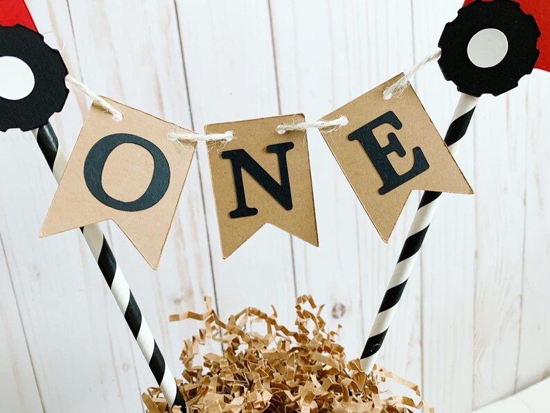May include: A brown paper banner with the word "ONE" in black letters, attached to black and white striped straws with red and black tractor wheels on top.