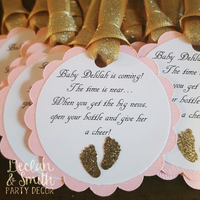 May include: Pink and white favor tags with gold glitter accents. The tags feature the text "Baby Delilah is coming! The time is near... When you get the big news, open your bottle and give her a cheer!" and two gold glitter footprints.