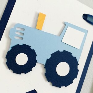Light blue tractor first birthday ONE banner. The base pennant flags are navy blue. The letters are light blue and outlined in navy blue. The tractors have closed cabs and are light blue with navy blue tires, white wheels, white window and light orange exhaust. The tractor banner is strung on navy satin ribbon.