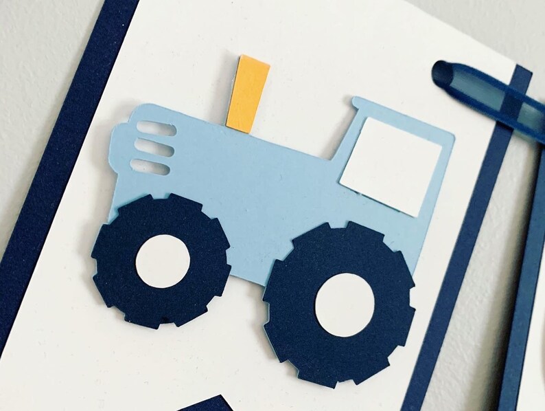 Light blue tractor first birthday ONE banner. The base pennant flags are navy blue. The letters are light blue and outlined in navy blue. The tractors have closed cabs and are light blue with navy blue tires, white wheels, white window and light orange exhaust. The tractor banner is strung on navy satin ribbon.