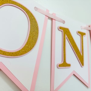 May include: Pink and gold glitter banner with the word "ONE" on it. The banner is made of cardstock and has a pink ribbon attached to each end.