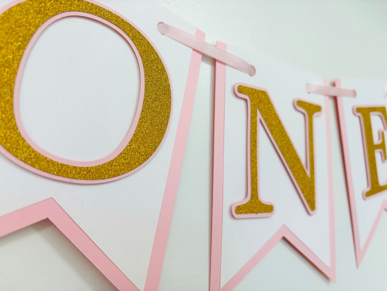 May include: Pink and gold glitter banner with the word "ONE" on it. The banner is made of cardstock and has a pink ribbon attached to each end.