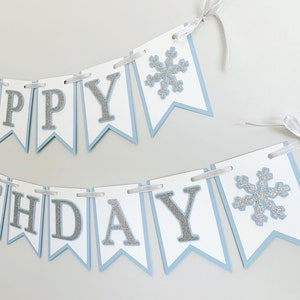 May include: A blue and white banner with silver glitter letters that reads "Happy Birthday". The banner has two silver glitter snowflakes.