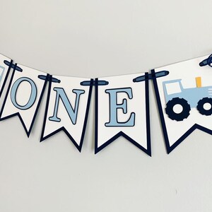 Light blue tractor first birthday ONE banner. The base pennant flags are navy blue. The letters are light blue and outlined in navy blue. The tractors have closed cabs and are light blue with navy blue tires, white wheels, white window and light orange exhaust. The tractor banner is strung on navy satin ribbon.