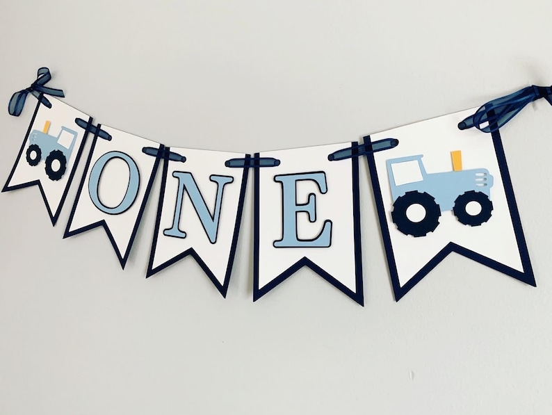 Light blue tractor first birthday ONE banner. The base pennant flags are navy blue. The letters are light blue and outlined in navy blue. The tractors have closed cabs and are light blue with navy blue tires, white wheels, white window and light orange exhaust. The tractor banner is strung on navy satin ribbon.