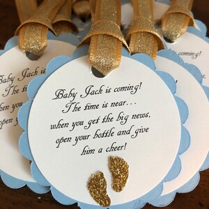 May include: Blue and white baby shower favor tags with gold glitter accents. The tags feature the text "Baby Jack is coming! The time is near... when you get the big news, open your bottle and give him a cheer!"