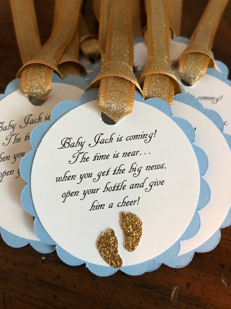 May include: Blue and white baby shower favor tags with gold glitter accents. The tags feature the text "Baby Jack is coming! The time is near... when you get the big news, open your bottle and give him a cheer!"