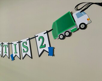 May include: A green and white garbage truck with a black ribbon attached to the top. The truck is pulling a banner with the words "IT IS 2" in green letters on white flags. There are two blue recycling bins hanging from the banner.