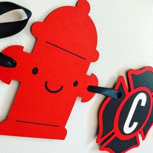 May include: Red fire hydrant cutout with a smiling face and black ribbon attached. A black and red fire department badge cutout with a white letter C in the centre is attached to the ribbon.