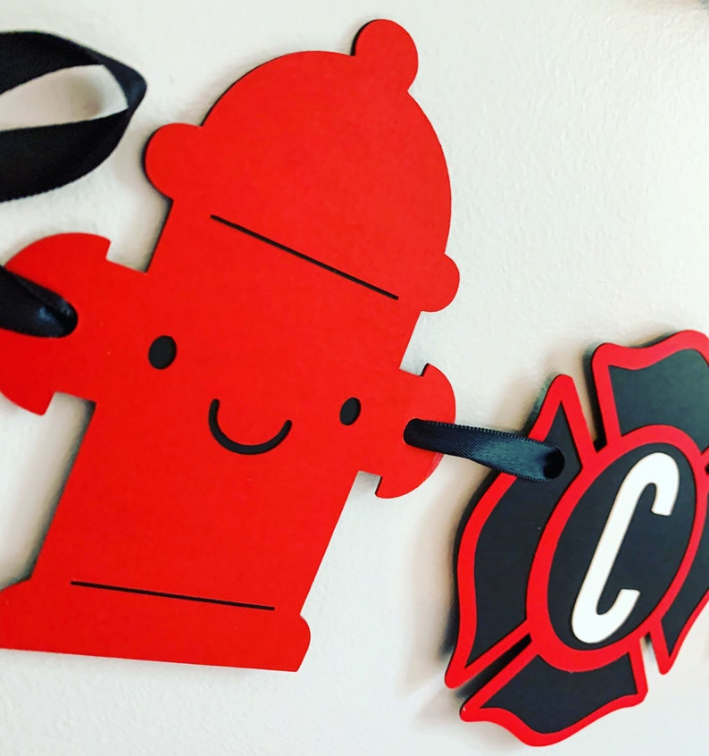 May include: Red fire hydrant cutout with a smiling face and black ribbon attached. A black and red fire department badge cutout with a white letter C in the centre is attached to the ribbon.