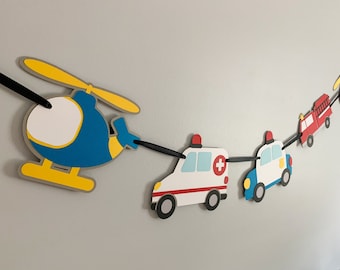 May include: A colourful paper banner featuring a blue helicopter, a white ambulance, a blue police car and a red fire engine. The banner is strung together with black ribbon.