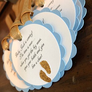 May include: Blue and white baby shower favor tags with gold glitter footprints and the text "Baby Jack is coming! The time is near... when you get the big news, open your bottle and give him a cheer!"