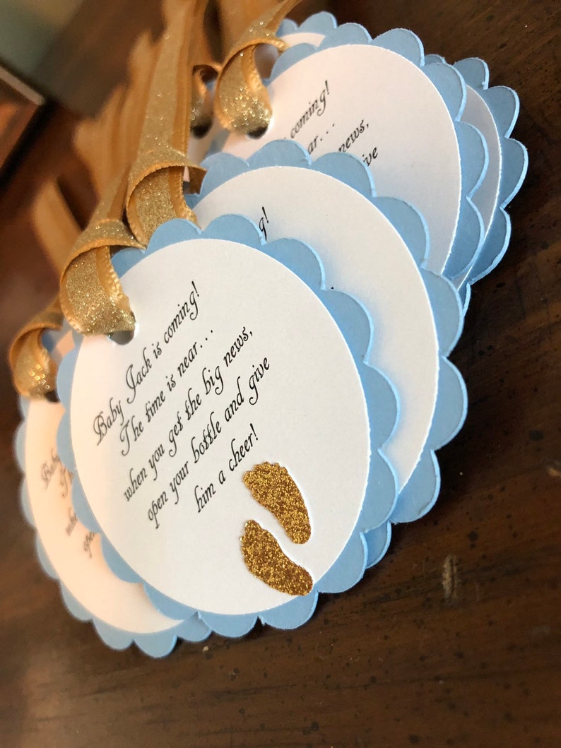 May include: Blue and white baby shower favor tags with gold glitter footprints and the text "Baby Jack is coming! The time is near... when you get the big news, open your bottle and give him a cheer!"