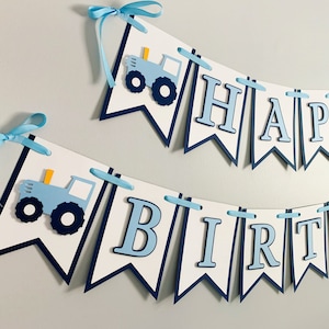 May include: A blue and white banner with the words "Happy Birthday" and two blue tractors. The banner is strung with blue ribbon.