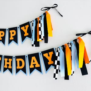 May include: A festive "HAPPY BIRTHDAY" banner with orange letters on black pennants, outlined in light blue. The banner is adorned with colorful ribbons in orange, yellow, black, and checkered patterns, perfect for a birthday celebration.