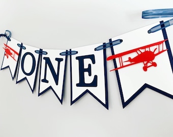 May include: A white and blue banner with the word "ONE" in navy blue letters. A red airplane silhouette is on the right side of the banner.