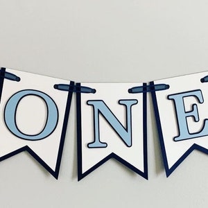 Light blue tractor first birthday ONE banner. The base pennant flags are navy blue. The letters are light blue and outlined in navy blue. The tractors have closed cabs and are light blue with navy blue tires, white wheels, white window and light orange exhaust. The tractor banner is strung on navy satin ribbon.