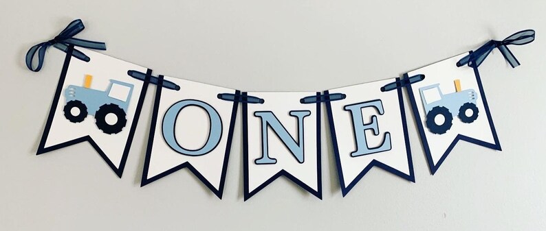 Light blue tractor first birthday ONE banner. The base pennant flags are navy blue. The letters are light blue and outlined in navy blue. The tractors have closed cabs and are light blue with navy blue tires, white wheels, white window and light orange exhaust. The tractor banner is strung on navy satin ribbon.
