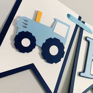 May include: Blue and white banner featuring a blue tractor and the letter "I". The tractor has a yellow chimney and white wheels. The banner is decorated with blue ribbon.
