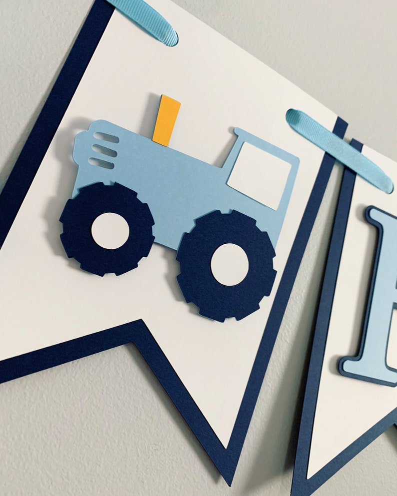 May include: Blue and white banner featuring a blue tractor and the letter "I". The tractor has a yellow chimney and white wheels. The banner is decorated with blue ribbon.