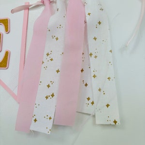 May include: Pink and white fabric banner with gold stars. The banner is made of strips of fabric with frayed edges and is tied together with pink ribbon.