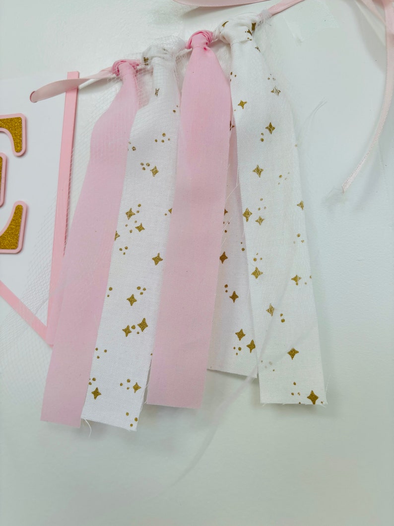 May include: Pink and white fabric banner with gold stars. The banner is made of strips of fabric with frayed edges and is tied together with pink ribbon.