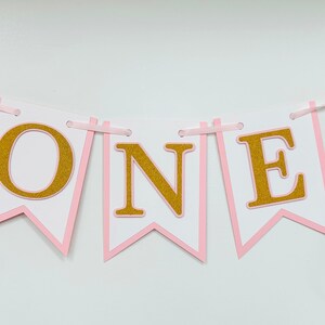 May include: Pink and gold banner with the word "ONE" in gold glitter letters. The banner has pink and white ribbon streamers.