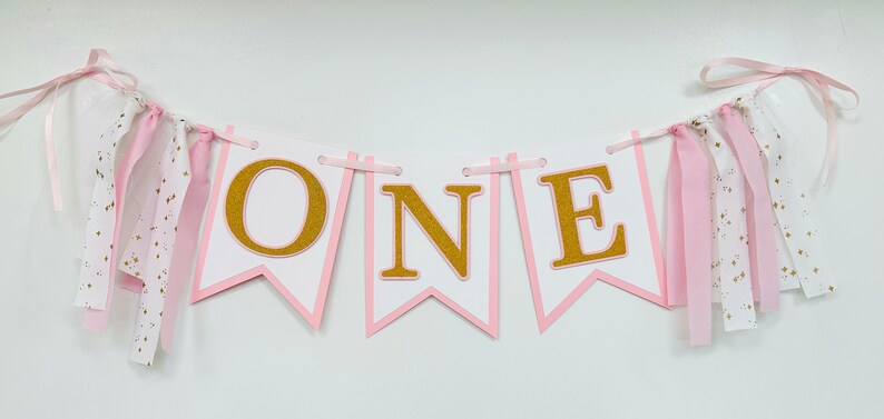 May include: Pink and gold banner with the word "ONE" in gold glitter letters. The banner has pink and white ribbon streamers.