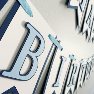 May include: A blue and white paper banner with the words "Happy Birthday" in blue letters. The banner is strung together with blue ribbon.