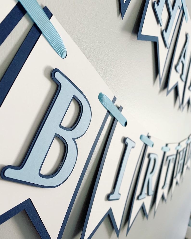 May include: A blue and white paper banner with the words "Happy Birthday" in blue letters. The banner is strung together with blue ribbon.