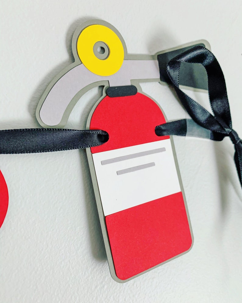 May include: A red and grey fire extinguisher paper craft with a yellow nozzle and black ribbon. The fire extinguisher has a white label with grey lines.