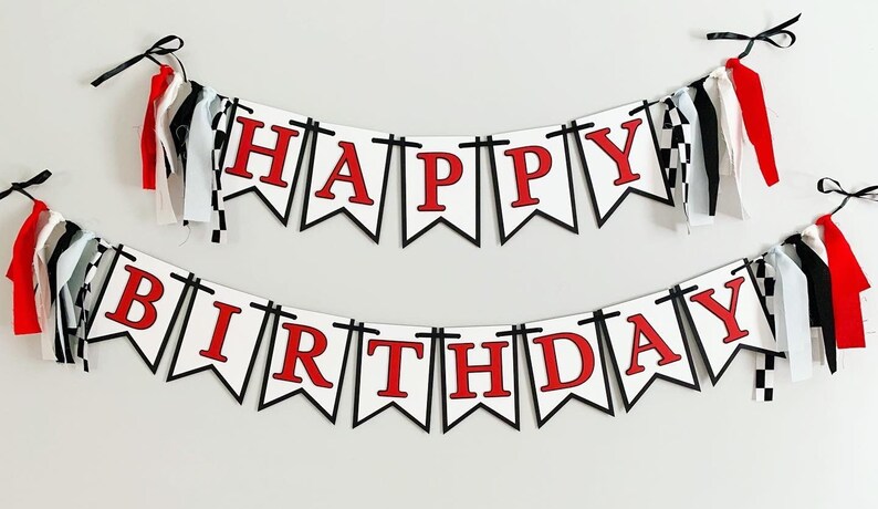 Op de afbeelding: Een wit-zwarte banner met rode letters die "Happy Birthday" lezen. De banner is versierd met zwart-wit geblokt lint en rode, witte en zwarte kwastjes.