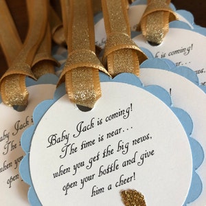 May include: Blue and white baby shower favor tags with gold glitter ribbon and gold glitter footprints. The tags read "Baby Jack is coming! The time is near... when you get the big news, open your bottle and give him a cheer!"